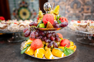 A vibrant fruit platter featuring grapes, watermelon, kiwi, and oranges on a decorative stand. The colorful fruits contrast beautifully against a blurred background of assorted desserts
