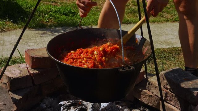 A traditional Romanian cooking moment where tomato puree is poured into a cauldron over a wood fire. This rustic outdoor scene captures the making of authentic zacusca, a flavorful spread.