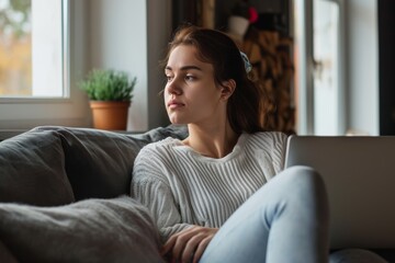 Obraz premium Young woman sitting on a couch indoors, distracted from work while gazing thoughtfully out the window during the afternoon