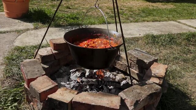 A traditional Romanian cooking moment where tomato puree is poured into a cauldron over a wood fire. This rustic outdoor scene captures the making of authentic zacusca, a flavorful spread