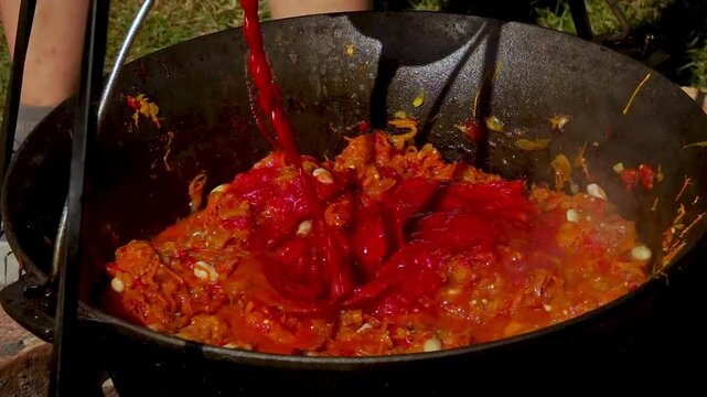 A traditional Romanian cooking moment where tomato puree is poured into a cauldron over a wood fire. This rustic outdoor scene captures the making of authentic zacusca, a flavorful spread