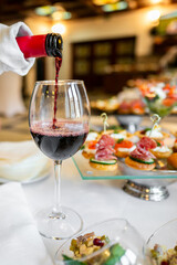 A close-up of a hand pouring red wine into a glass, with a variety of appetizers in the background. The scene captures the elegance and enjoyment of a wine tasting experience