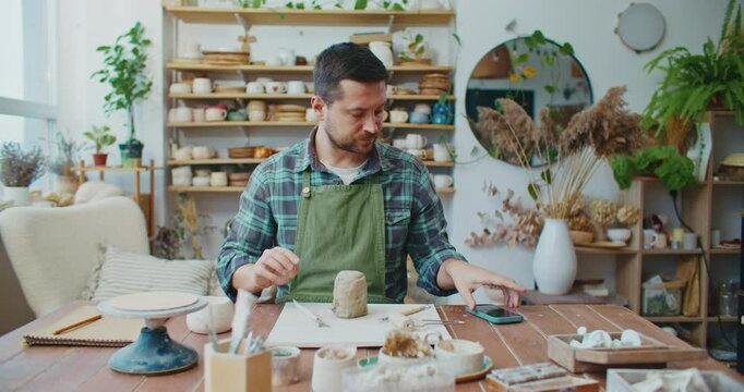 Focused Caucasian male peeling piece of clay with instrument. Potter preparing material for new creation. Getting distracted by mobile phone. Picking up smartphone. Searching for solution online.