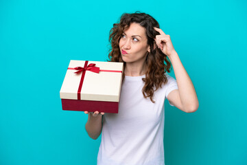 Young caucasian woman holding a gift isolated on blue background having doubts and with confuse face expression