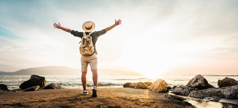 Happy man with backpack standing with arms up at the beach - Delightful tourist enjoying summer vacation by the seaside - Traveling life style and well being concept