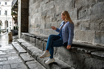 Beautiful young woman wearing jeans and jacket sitting on stone wall against background of old building doing something on smartphone on autumn day. Break in sightseeing in old town. Bergamo, Italy.