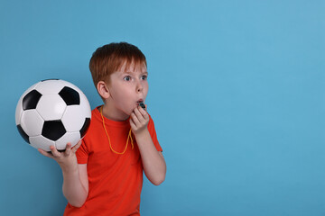 Little boy with soccer ball blowing whistle on light blue background, space for text