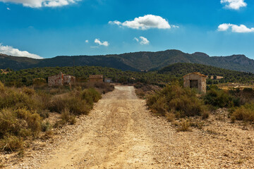 Gravel road with some sheds in spanish countryside with mountains and vegetation under blue cloudy...