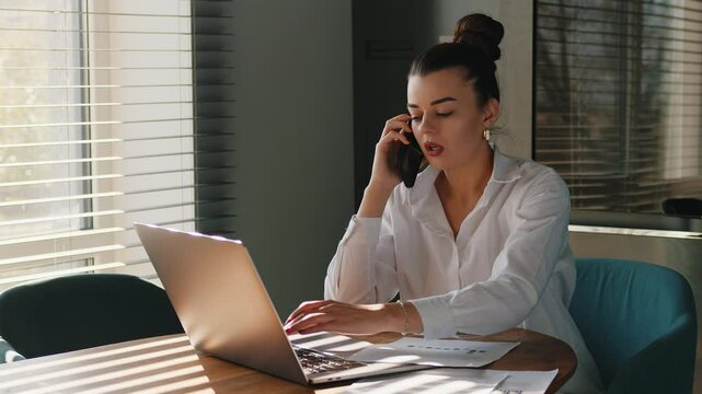 Young busy business woman manager using laptop and mobile cell phone devices in home office. Professional female executive holding smartphone, working on laptop sitting at desk.