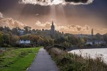 Serene path winding alongside the water, framed by lush greenery and the stunning church tower...