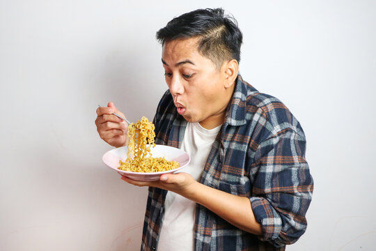 Young handsome trendy asian man eating yummy hot and spicy instant noodle using fork isolated on white background. Asian guy servile end of the month with cheap food.