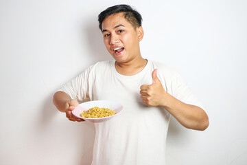 Young Asian man in a white t-shirt happily pointing at a bowl of instant noodles. Studio shot on a plain background, showcasing his excited expression and casual fashion