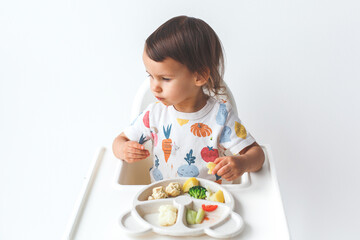 a little cute girl baby 1.5 years old in a bright T-shirt sits in a white high chair against a white wall in the kitchen and eats with her hands.