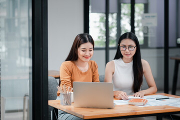 Two young women working together at a desk with a laptop and papers in a modern office setting.