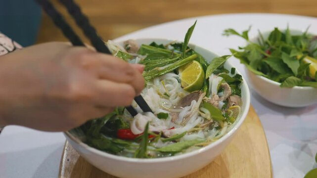 Female hand stirring vietnamese beef noodle soup pho bo with black chopsticks.