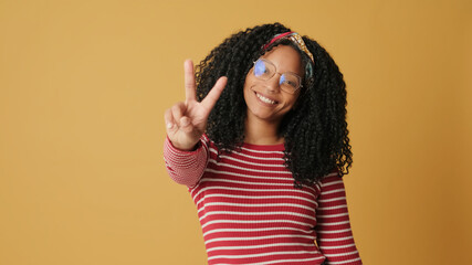 Young smiling woman with curly hair wearing glasses wearing striped sweater looking at camera showing fingers victory sign gesture while standing on yellow background in studio