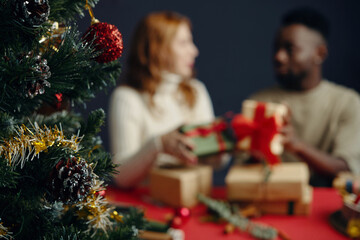 Two people enjoying Christmas celebration, exchanging gifts with smiles, sitting at decorated table with Christmas tree in foreground. Holiday season creating joyful atmosphere