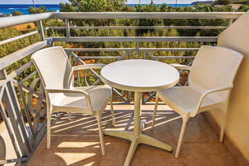 Plastic chairs and round table on balcony hotel with Mediterranean view, surrounded by green trees in warm sunlight. Greece.