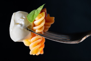 A close-up of a silver fork with fusilli pasta in tomato sauce with a mozzarella ball and  basil...
