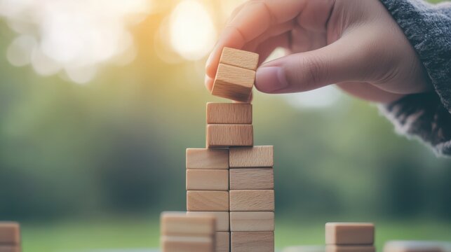 Retro toned image of man building a tower of blank wooden blocks outdoors against green with sparkling bokeh of sunlight.
