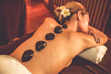 Hot stone massage at spa salon in luxury resort with warm candle light, blissful woman customer enjoying spa basalt stone massage glide over body with soothing warmth. Quiescent