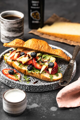 Food photography, a grey stone plate with a croissant sandwich on it. The croissant is filled with navy blue berries and slices of strawberries, apple, mozzarella cheese, and arugula salad. 