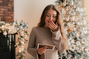 Side view of charming woman reading Christmas greeting card on background of Xmas tree at cozy...