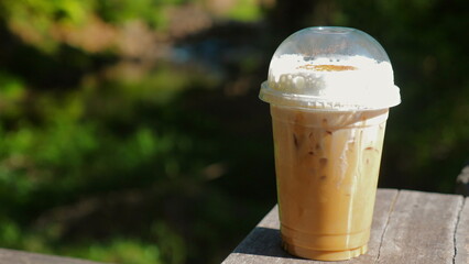 Iced coffee in plastic cup with whipped cream on wooden table, outdoor background on sunny day. Refreshment and Leisure