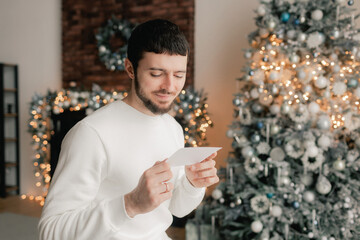 Side view of man reading Christmas greeting card on background of Xmas tree at cozy living room...