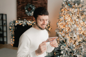 Side view of man reading Christmas greeting card on background of Xmas tree at cozy living room with festive interior. Surprised lady pleasure reading letter at home. Part of a series