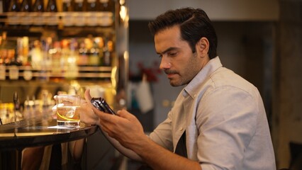 Smart man drinking cocktail in Old fashioned waiting friends or couple while chatting on smartphone to wait at counter bar with bartender preparing drinks at nightclub Friday meeting night. Vinosity.