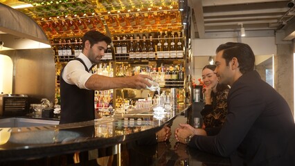 Focusing smiling smart bartender serving gin fizz or tonic soda at luxurious bar, engaging lively conversation with VIP customer couple at back view in atmosphere at lighting warm nightclub. Vinosity.