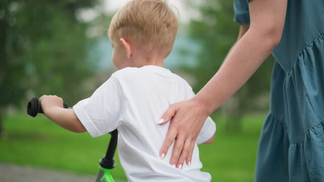 A woman with painted nails supports her young son who is learning how to ride a bicycle, the boy uses his leg for balance as she lets him move forward independently