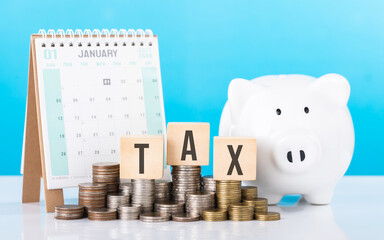Wooden block labeled 'TAX' placed next to a stack of coins, a calendar, and a piggy bank. Image...