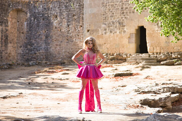 Drag queen, blonde, attractive, with crown, wearing pink dress and heeled boots, posing facing the camera with her hands on her hips, in the middle of a castle courtyard. Concept pride, identity.