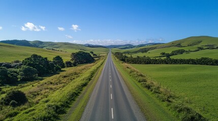 A straight, paved road winds through a countryside landscape. Green hills dotted with trees and bushes stretch out under a bright blue sky. It's a summer scene.