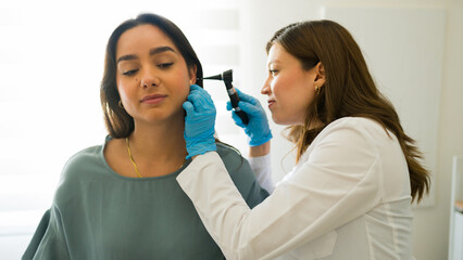 Female doctor wearing gloves is using an otoscope to examine the ear of a young woman in a medical...