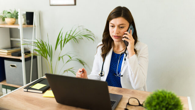 Female doctor is working in her office, talking on the phone and taking notes on her laptop