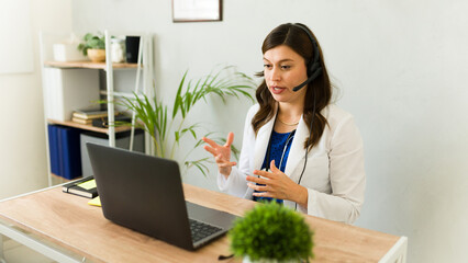 Female doctor in white coat using headset for video call with patient on laptop at clinic