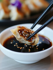close-up of a dumpling being dipped into a bowl of soy sauce with sesame seeds, held by black chopsticks. The background appears blurred, highlighting the dumpling and sauce