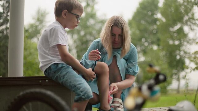 A mother squats as she carefully cleans her son's bleeding leg with a tissue while the boy holds his pant leg up