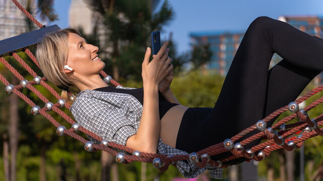 Smiling blonde young woman lies in a rope hammock and listens to music through wireless headphones on a sunny day in the park. Concept of recreation