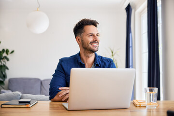 Smiling thoughtful man working on his laptop from home looking away