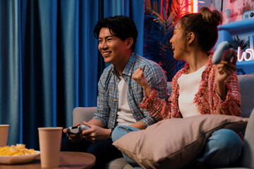 Couple gamer raising fist up holding joysticks be winner together complete level sitting sofa playing video game with front snack and drinks at neon light color living room at modern home. Infobahn.