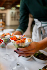 A close-up of hands holding a glass bowl filled with a fresh salad, featuring cubed cheese and sliced vegetables. The blurred background suggests a dining setting, emphasizing the vibrant colors