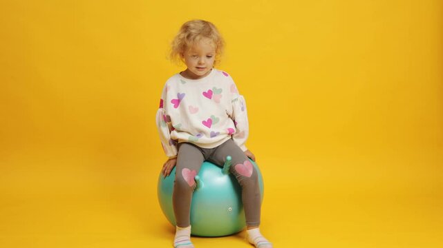 Happy child jumping on bouncing ball. Toddler wavy haired blonde girl playing on gymnastic ball kid having fun with excitement showing like gesture isolated over yellow background