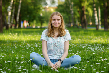 Fototapeta premium A young woman sitting cross-legged on a grassy field filled with daisies. She has long blonde hair and is wearing a light floral shirt and blue jeans. The background features lush green trees