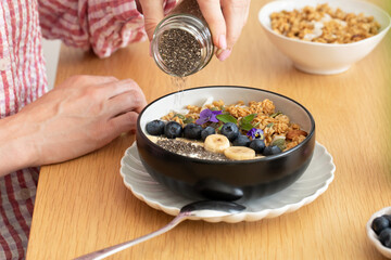 A woman pours chia seeds on granola and Greek yogurt. Healthy breakfast.