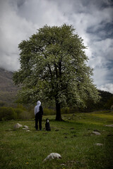 A girl and a dog are standing under a tree. A green tree. Clouds in the sky.