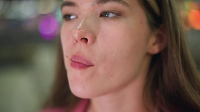 Close-up of a woman licking her pink lips as if she has something in her mouth spots are seen on her face, with colored blurred lights blinking in the background, and unclear object in behind her